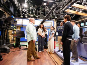 Group of UM students listening attentively to a ESPN employee talking, in a recording studio with a bunch of lights rigged above.